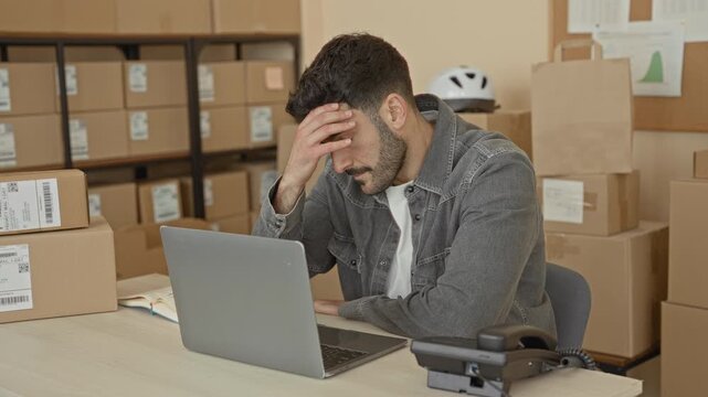 Man at laptop touches chin and forehead among stacked shipping boxes and labels in building; small business concern.