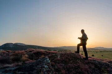 Obraz premium silhouette of a man photographing in the mountains, A person is silhouetted against the setting sun while walking along a rocky, flower-covered ridge overlooking a vast landscape. Sardinia Italy 