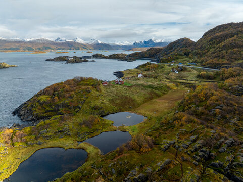 Aerial view of Hamn i Senja featuring rocky islands, small ponds, and red houses against a backdrop of snow-capped mountains in Skaland, Troms, Norway.