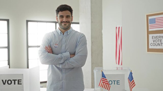 Man with arms crossed wearing an i voted sticker beside a ballot box and small us flags in a polling building; pride civic duty.