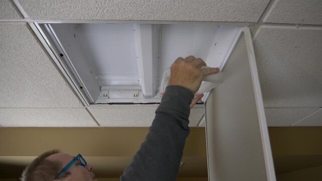 Old middle aged man changing a u-shaped light bulb in a ceiling light fixture in a drop ceiling in a basement