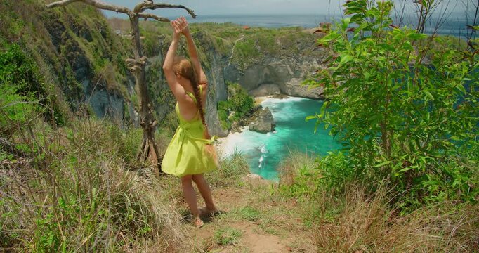 Smiling young woman in summer dress enjoying ocean view and dancing along steep cliff path with scenic view background