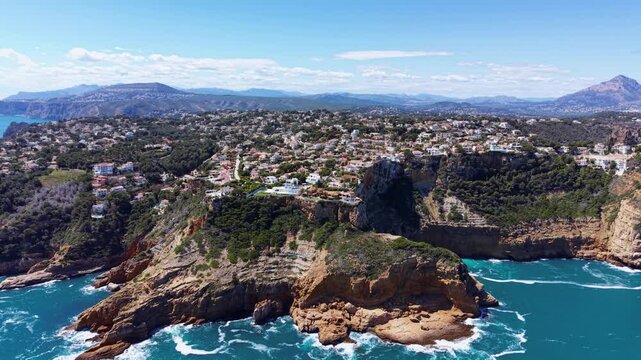 Aerial view of Cap de la Nau, Javea, Spain, featuring luxury villas perched on dramatic rocky cliffs above the turquoise Mediterranean Sea with mountains in background