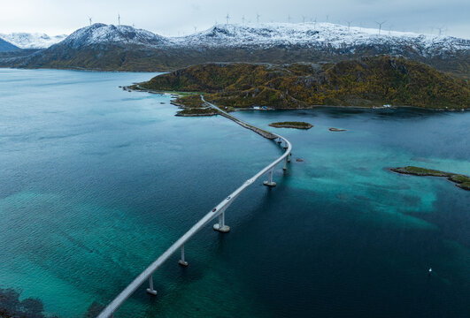Aerial view of the Sommaroy Bridge stretching across turquoise coastal waters with snow-capped mountains and wind turbines in the background Sommaroy, Troms, Norway.