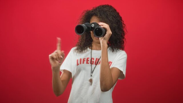 Woman lifeguard in lifeguard tshirt holds binoculars to her eyes and points index finger against a red studio backdrop; alert duty.
