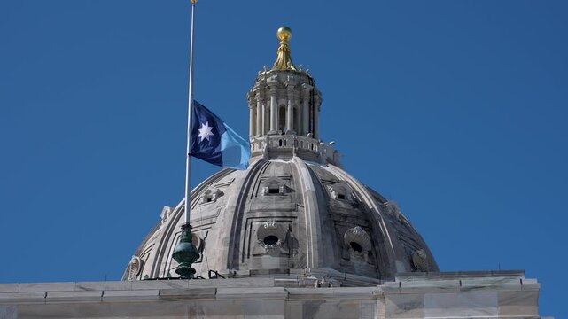 Minnesota state flag waves over State Capitol building in St Paul under bright sun and clear blue sky on a warm day