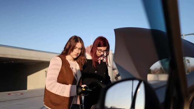 View from inside car two women explore engine under open hood using tools carefully testing ideas and trying to bring vehicle back to life during unexpected breakdown