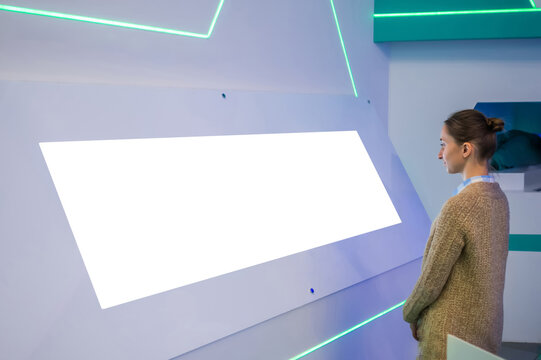Mockup image: woman looking at blank large white interactive wall display at modern technology exhibition, museum, trade show. Mock up, white screen, copyspace, template, education concept