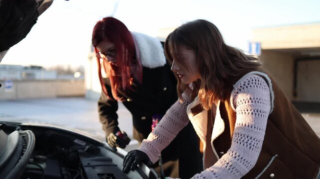 Two women try to fix broken car under open hood using tools focusing on engine problem working together outdoors in parking area learning repair without professional help