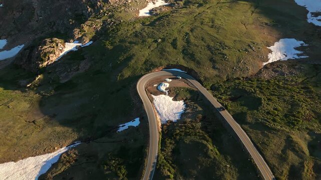 Beartooth Highway in Montana curves through alpine terrain with scattered snow patches and green tundra. Cars navigate the scenic switchback during warm evening light.