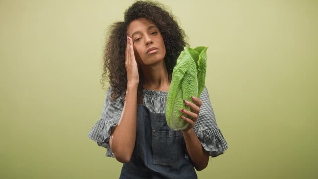Woman in denim apron holding romaine lettuce with hand to cheek and eyes closed on a green studio backdrop, curly hair visible; fatigue.