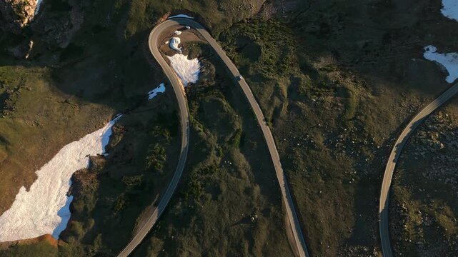 Aerial drone footage showcases dramatic serpentine mountain highway winding through rugged alpine terrain with patches of melting snow scattered across green mossy hillsides during warm golden hour.