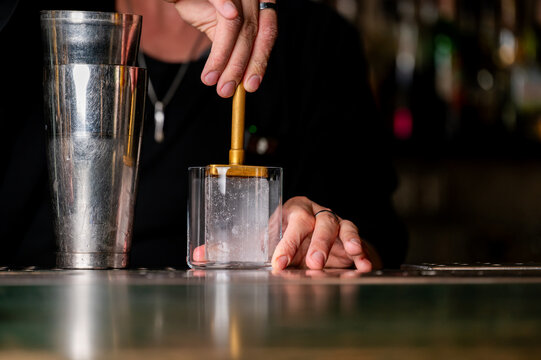 Close-up of a bartender using a brass brander to sear a logo onto a large clear ice cube in a rocks glass, next to a stainless steel cocktail shaker on a bar counter.