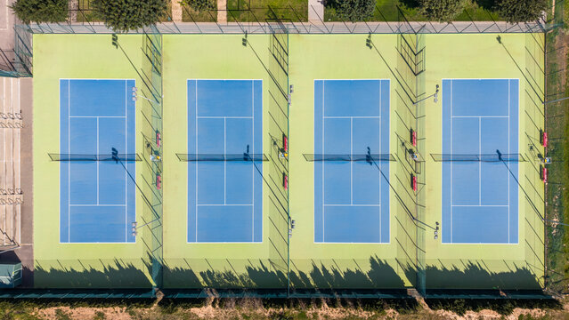 Overhead aerial view of four parallel tennis courts with bright blue playing surfaces and green surrounds, casting long shadows, with players visible on the courts