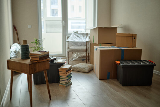 Wide shot of packed moving boxes, storage bins, books, chair, and decor filling empty apartment, suggesting relocation and home setup. Ideal for moving services, real estate, downsizing