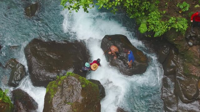 Top down drone shot of three adult men carefully passing a young boy hand to hand while crossing a shallow rocky river in tropical forest, teamwork and protection concept.