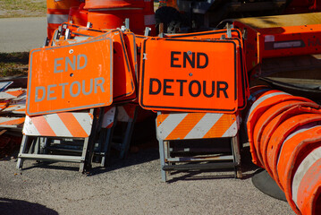Naklejka premium Bright orange End Detour signs and stacked traffic cones rest on pavement at a road construction site, conveying traffic control, temporary closure, and urban maintenance work in daylight. 