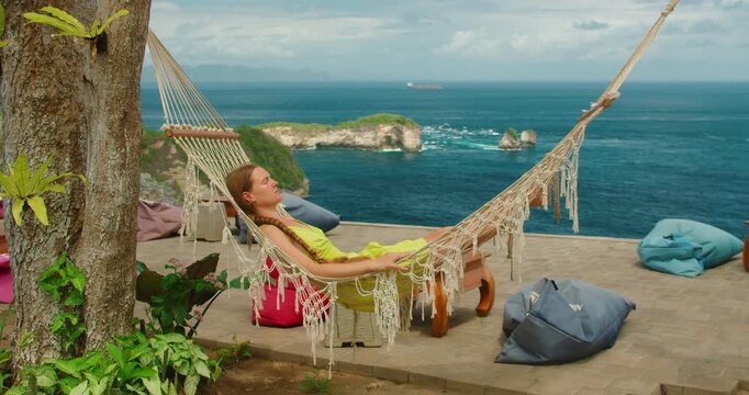 Relaxed woman resting in a white macrame hammock with a backdrop of tropical cliffs and ocean