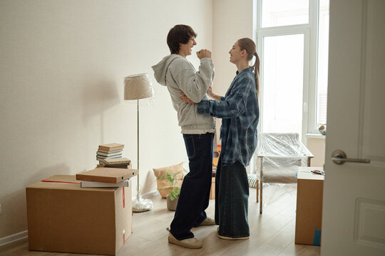 Young adult couple embracing and celebrating in new apartment among moving boxes, marking relocation and fresh start. Ideal for real estate, mortgage, rental, moving service marketing