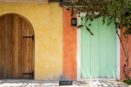 Two contrasting doors, one arched wooden and one mint green shutter, set in yellow and orange stucco walls with foliage, showcasing traditional Mediterranean architectural detail in daylight.