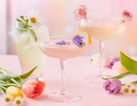 Spring cocktails with floral garnishes are placed on a pastel table. Colorful tulips and daisies frame the drinks, creating a fresh and festive mood. The light-colored beverages look inviting for a ce