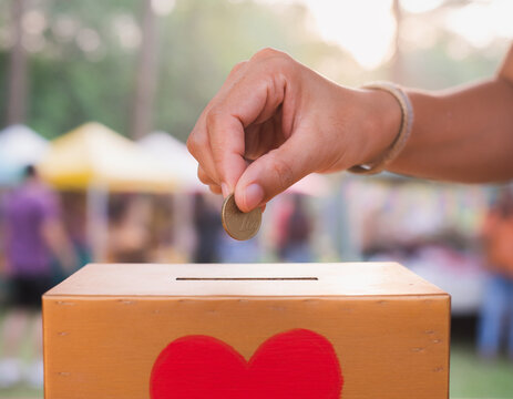 A close-up of a hand placing a coin into a wooden donation box with a red heart symbol at an outdoor event. This image is ideal for illustrating charity, community support, or fundraising concepts.