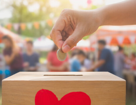 A close-up of a hand placing a coin into a wooden donation box with a red heart symbol at an outdoor event. This image is ideal for illustrating charity, community support, or fundraising concepts.