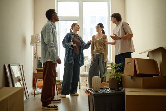 Multiethnic young adults discussing move-in plans among packed boxes in new apartment, planning relocation and shared housing. Wide shot, useful for rental, real estate, moving service marketing