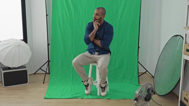 Latin man seated on stool touching chin in studio with green screen and softbox reflector fan; contemplation pondering.