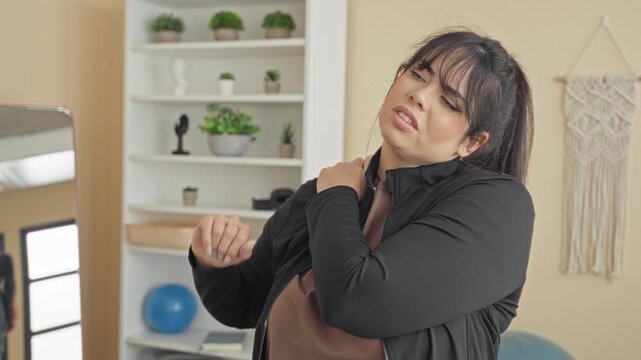 Plus size woman rubbing shoulder and wincing beside blue exercise ball and mirror in yoga studio; self care discomfort.