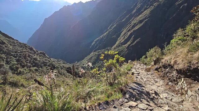 Inca Trail, Peru - 25 June 2024. Early sun lights a stone path above a deep valley, with grasses and shrubs in the foreground and shadowed mountain ridges beyond.