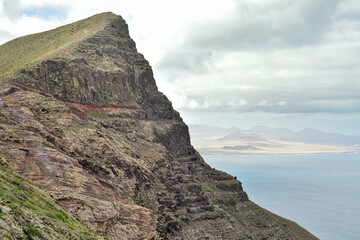Steep volcanic mountain cliff with visible sedimentary layers overlooking the ocean in Lanzarote. Arid coastal landscape with distant hills under a cloudy sky. © dsbedarieva