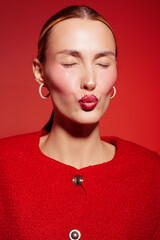 Close-up of a woman with closed eyes and red lipstick blowing a kiss, wearing red clothes and hoop...