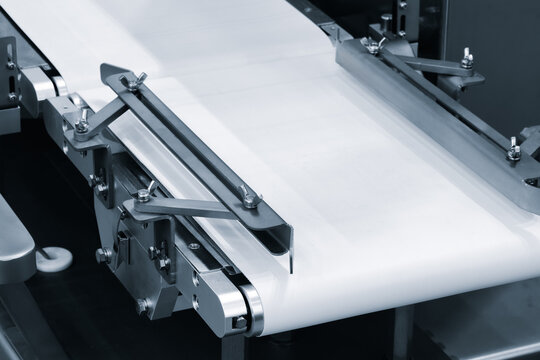 Close-up of industrial conveyor belt with metal guides and rollers. Smooth white belt moves steadily through machinery. Metal parts gleam under bright light