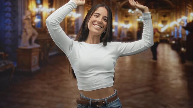 Woman smiling raises arms showing midriff in ornate museum building with chandeliers and statues; joy carefree celebration.