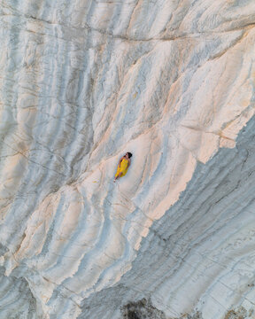 Aerial view of a woman in a yellow dress lying on the white marl cliffs of Riserva Naturale di Punta Bianca in Palma di Montechiaro, Sicily, Italy.