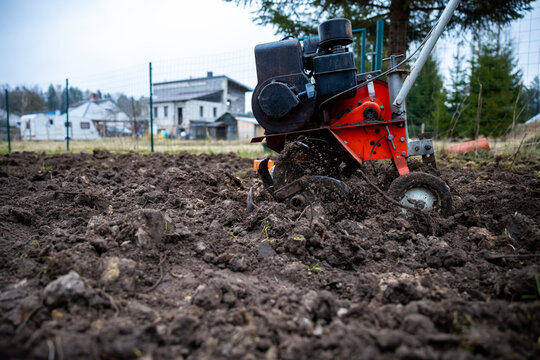 Gas powered rotary tiller bites into dark cloddy soil, tines fling earth in a fenced backyard near modest houses and outbuildings under overcast daylight.