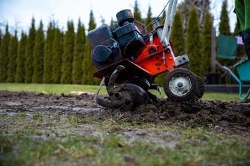 Obraz premium Ground level view shows metal tines churning earth by a mud splattered wheel. Worn paint, exposed belts, and compact engine mark an older machine with tools beyond.