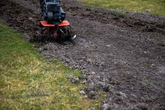A motorized tiller with an orange deck cuts compact soil into a straight row between short grass. Exposed tines churn earth in overcast light, tight low composition.