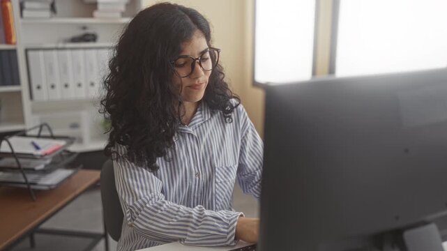 Young arab woman wearing glasses and striped shirt with hand on cheek at office building desk working on a desktop computer with a pained expression; discomfort toothache.