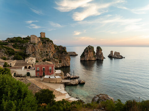 Aerial view of the historic Tonnara di Scopello with its red buildings and ancient towers overlooking the sea stacks and turquoise waters at sunset in Scopello, Sicily, Italy.