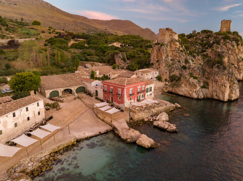 Aerial view of the historic Tonnara di Scopello with its red building, stone warehouses, and ancient coastal towers on rocky cliffs overlooking the turquoise sea in Scopello, Sicily, Italy.