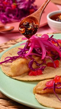 Hands pouring salsa macha over an Ensenada-style fish taco with a wooden spoon