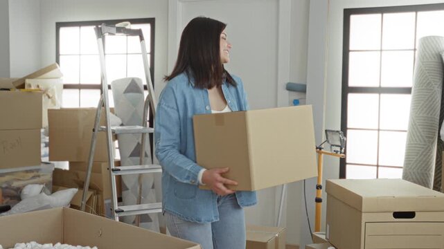Woman holding and lifting a cardboard moving box near ladder and stacked crates in building during relocation and packing; fresh start optimism.