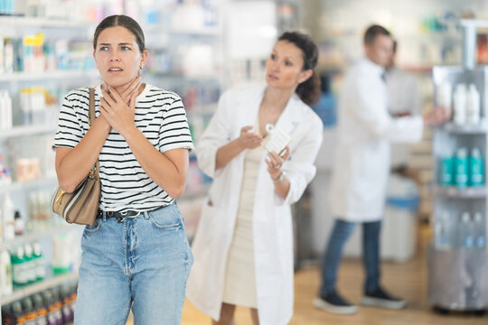 Young girl feeling pain standing with her back to female pharmacist offering remedy to her in drugstore