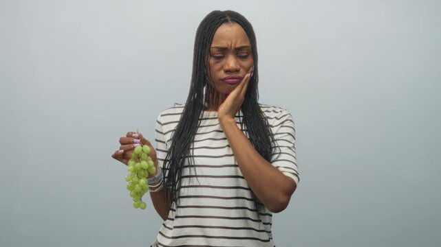 Black woman wearing striped shirt holding green grapes, touching cheek with hand for toothache in a studio; discomfort.