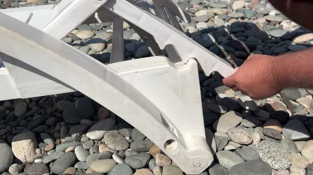 Horizontal 4K video. White man changes, sets height, tilt of backrest of plastic sun lounger, chaise longue, deck chair on pebble beach, sea coast. Male hand close up. Travel, relax, rest concept