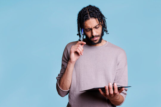 Pensive arab man thinking while holding clipboard and planning. Thoughtful young adult person in doubt reading checklist and touching face with pen with puzzled expression