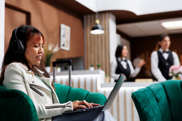 Smiling Asian tourist sitting at laptop listening to music in cozy hotel lounge area while on vacation. Woman guest relaxing in modern resort entrance waiting to book room