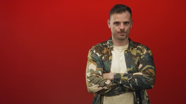 Man wearing camouflage army jacket and beige shirt, arms crossed with visible forearms, neutral gaze photographed in studio with red backdrop; confidence duty service.